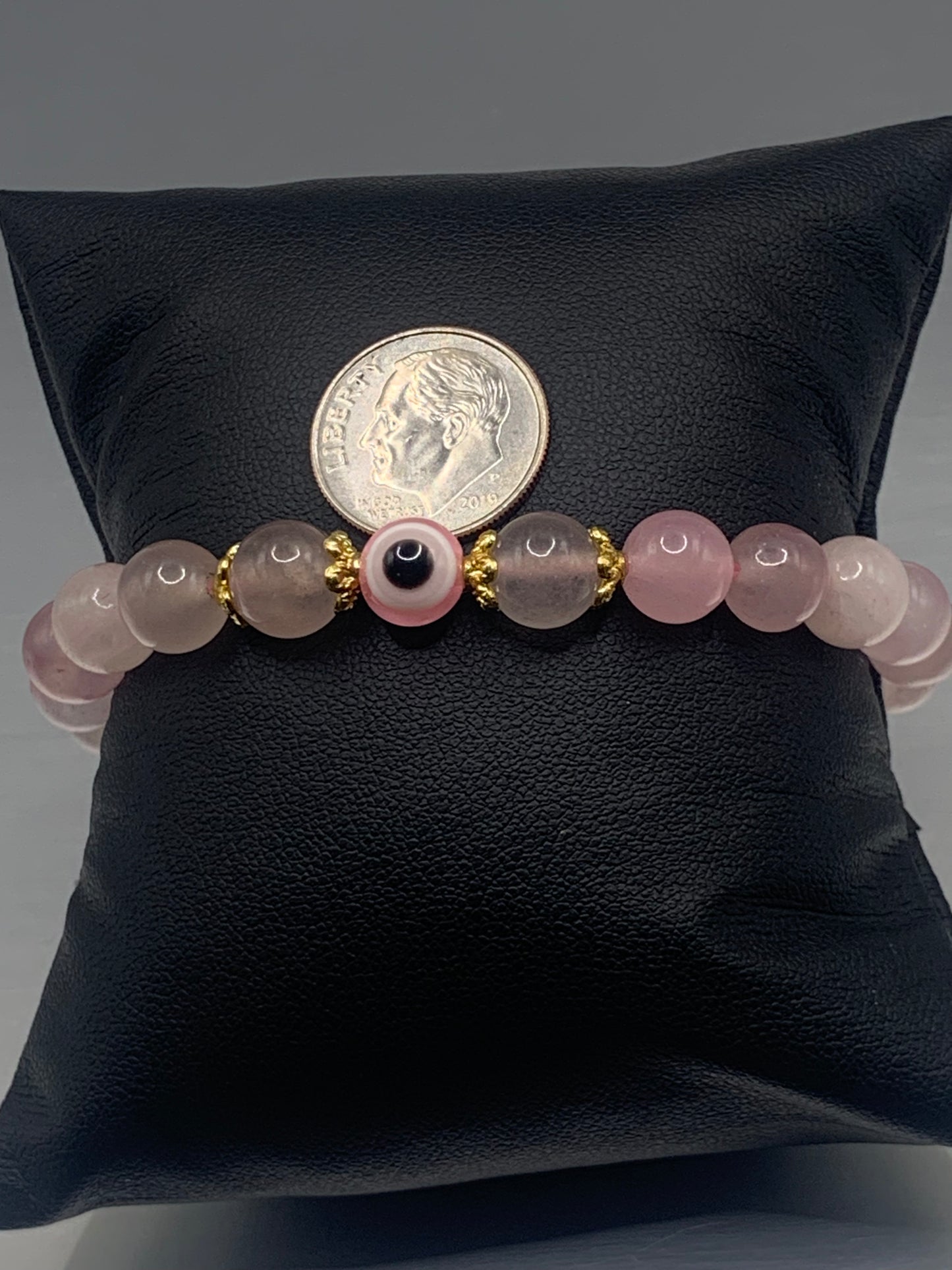 This bracelet features rose quartz (enhanced) beads with a resin evil eye as the focal bead. Each bead on either side of the evil eye have bead caps to draw attention to the evil eye bead. This picture is taken with the bracelet on a pillow with a dime to show a size ratio
