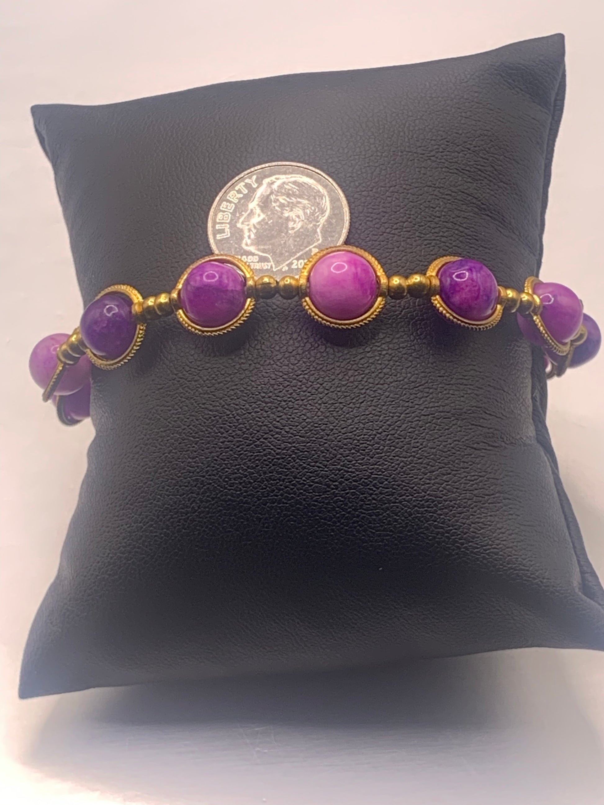 Purple charoite beaded bracelet with gold accents on a black cushion, next to a coin for scale.