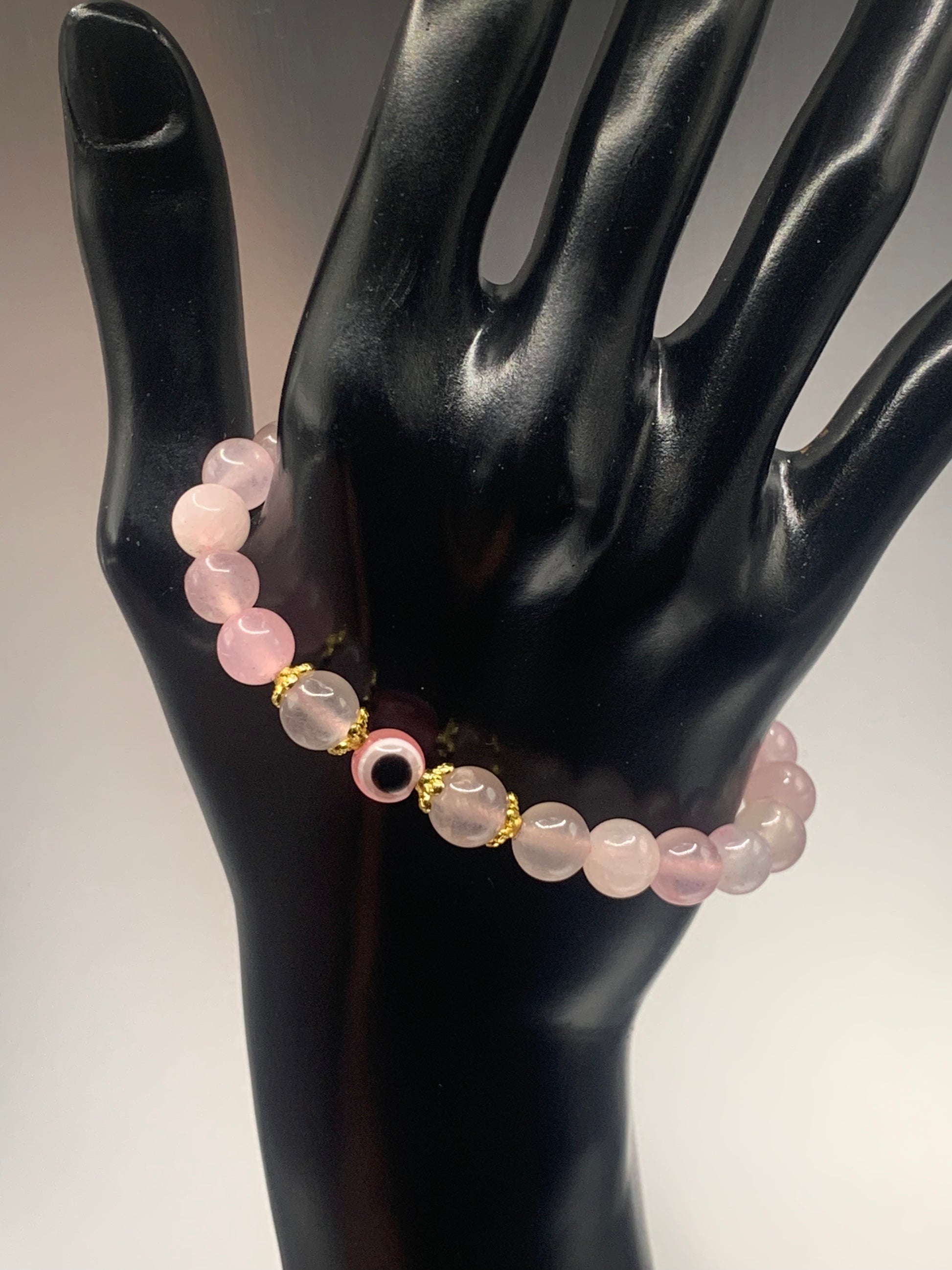 This bracelet features rose quartz (enhanced) beads with a resin evil eye as the focal bead. Each bead on either side of the evil eye have bead caps to draw attention to the evil eye bead. This picture is taken with the bracelet dangling on a hand