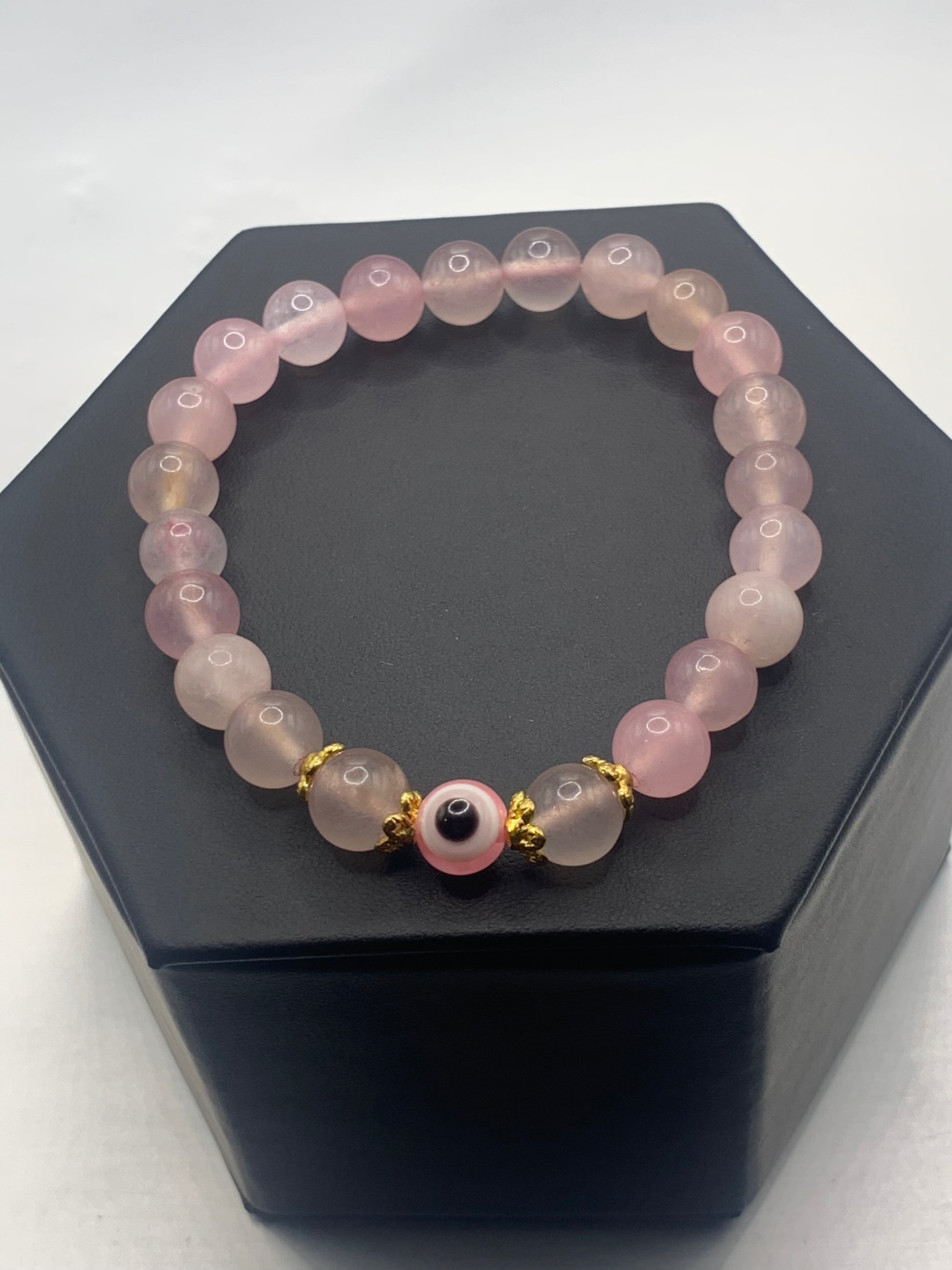 This bracelet features rose quartz (enhanced) beads with a resin evil eye as the focal bead. Each bead on either side of the evil eye have bead caps to draw attention to the evil eye bead. This picture is taken with the bracelet laying on a bracelet display 
