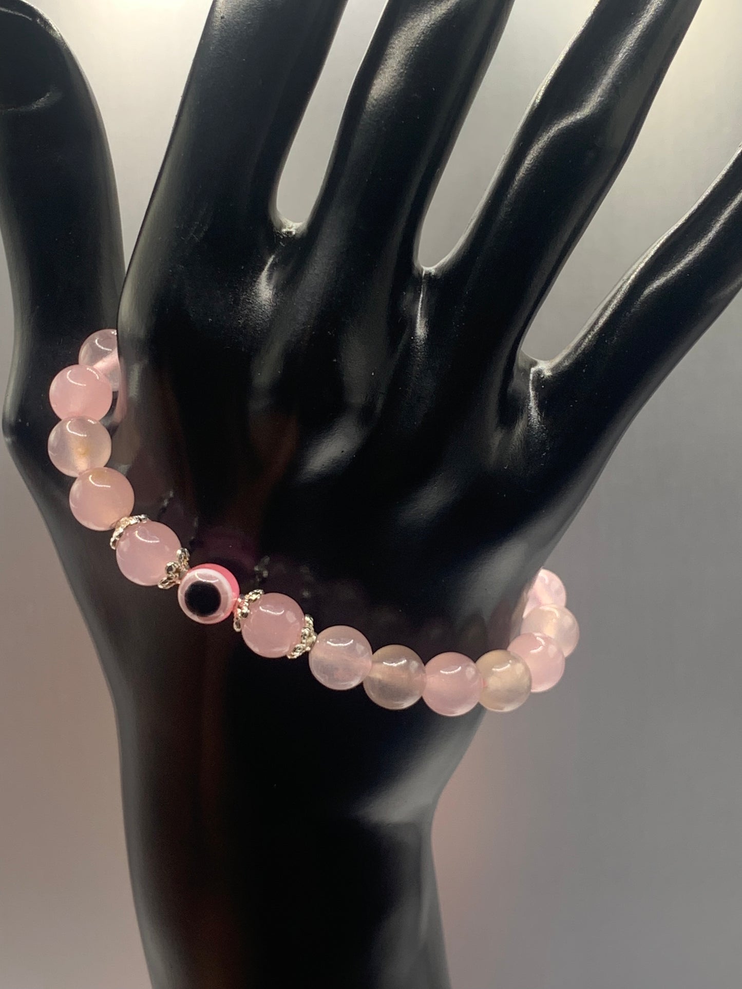 This bracelet features rose quartz (enhanced) beads with a resin evil eye as the focal bead. Each bead on either side of the evil eye have bead caps to draw attention to the evil eye bead. This picture is taken with the bracelet dangling on a hand
