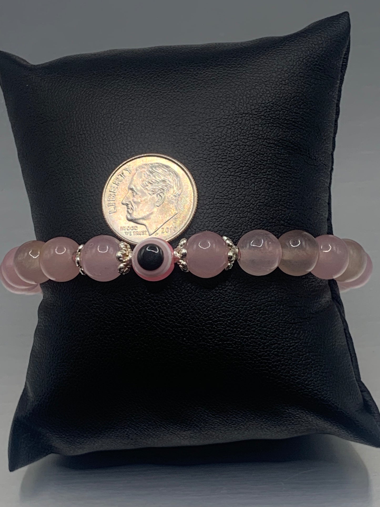 This bracelet features rose quartz (enhanced) beads with a resin evil eye as the focal bead. Each bead on either side of the evil eye have bead caps to draw attention to the evil eye bead. This picture is taken with the bracelet on a pillow with a dime to show a size ratio