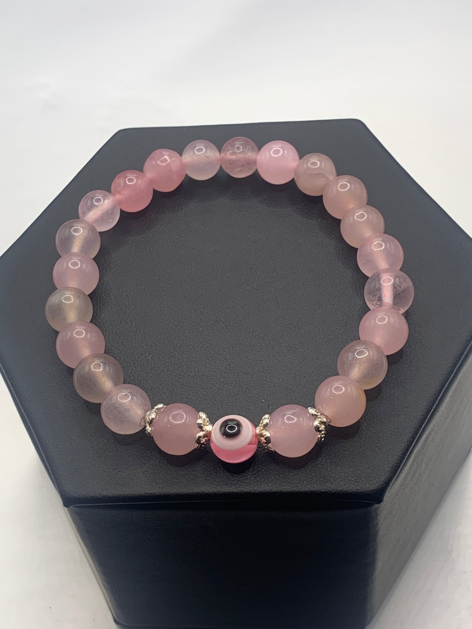 This bracelet features rose quartz (enhanced) beads with a resin evil eye as the focal bead. Each bead on either side of the evil eye have bead caps to draw attention to the evil eye bead. This picture is taken with the bracelet laying on a bracelet display 