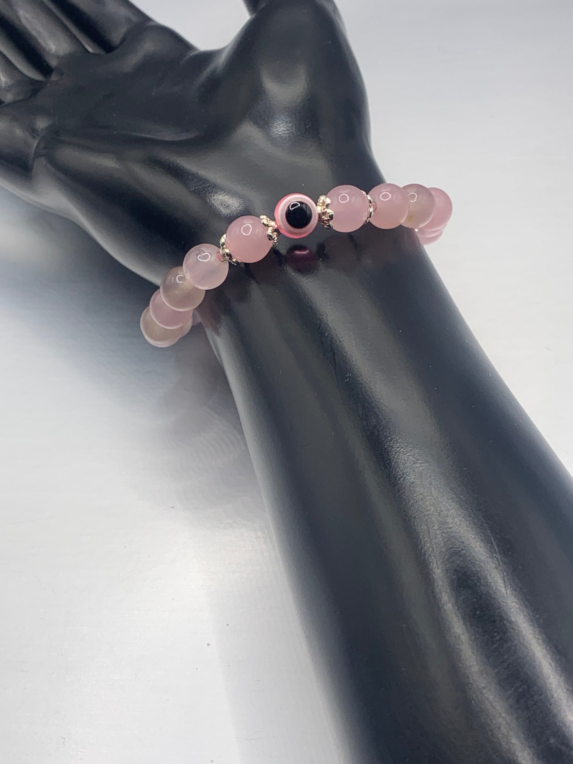 This bracelet features rose quartz (enhanced) beads with a resin evil eye as the focal bead. Each bead on either side of the evil eye have bead caps to draw attention to the evil eye bead. This picture is taken with the bracelet being worn on a wrist 
